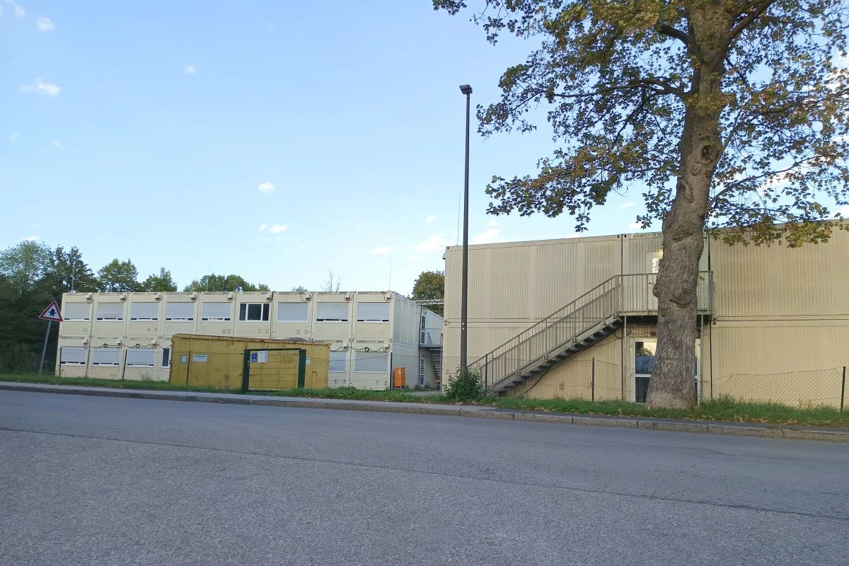 Container-Unterkunft mit heruntergelassenen Rolläden, quer davor verlaufender Straße und großem Baum (Foto: Peter Marwan)